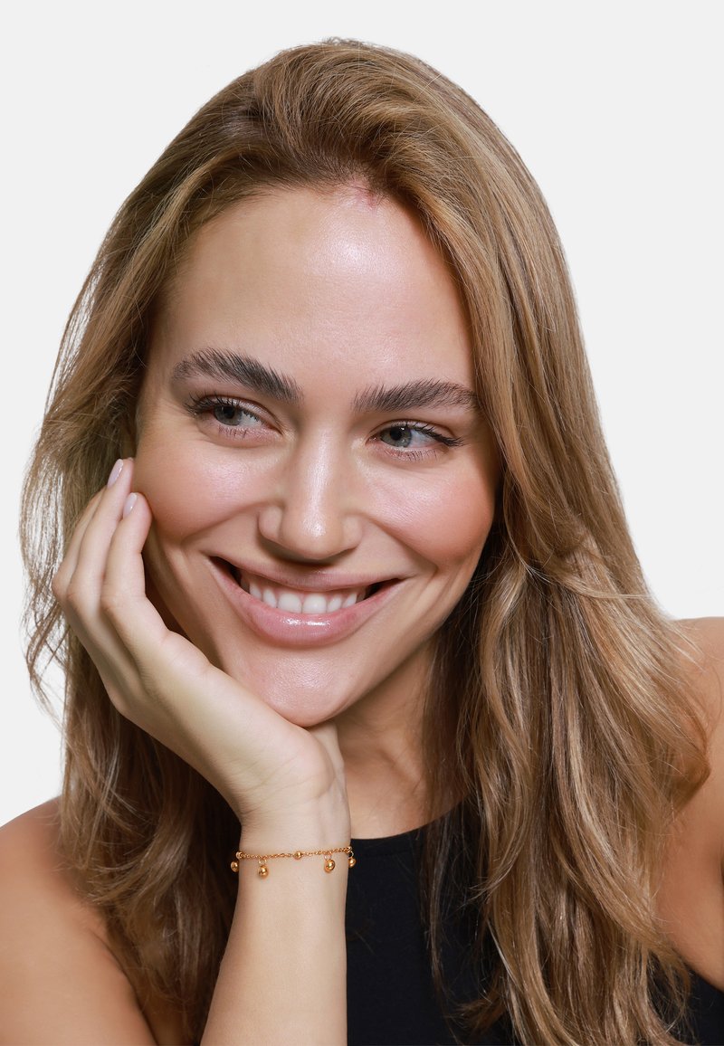 Bracciale d'oro con ciondoli a goccia appesi a un polso, indossato da una donna con lunghi capelli castano chiaro e ondulati e un sorriso dolce.