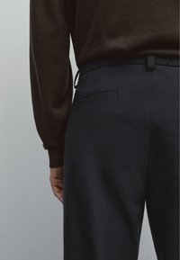 Pantaloni neri con una texture liscia, dotati di una cintura elastica, coulisse e una tasca laterale. Maglione marrone parzialmente visibile.