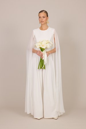 Femme en combinaison blanche à jambes larges avec des manches cape fluides tenant un bouquet de callas blanches noué avec un ruban sur un fond beige.
