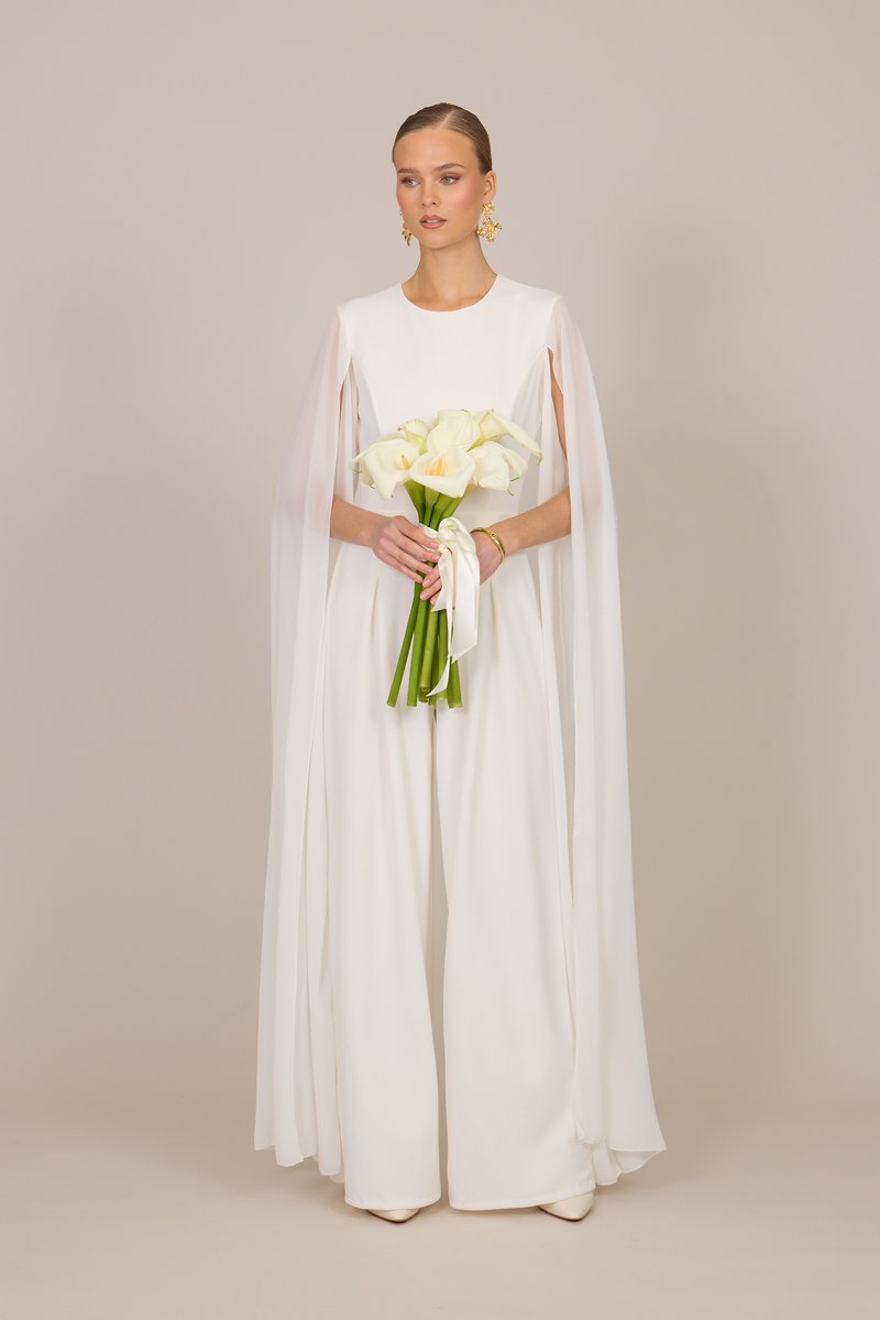 Femme en combinaison blanche à jambes larges avec des manches cape fluides tenant un bouquet de callas blanches noué avec un ruban sur un fond beige.