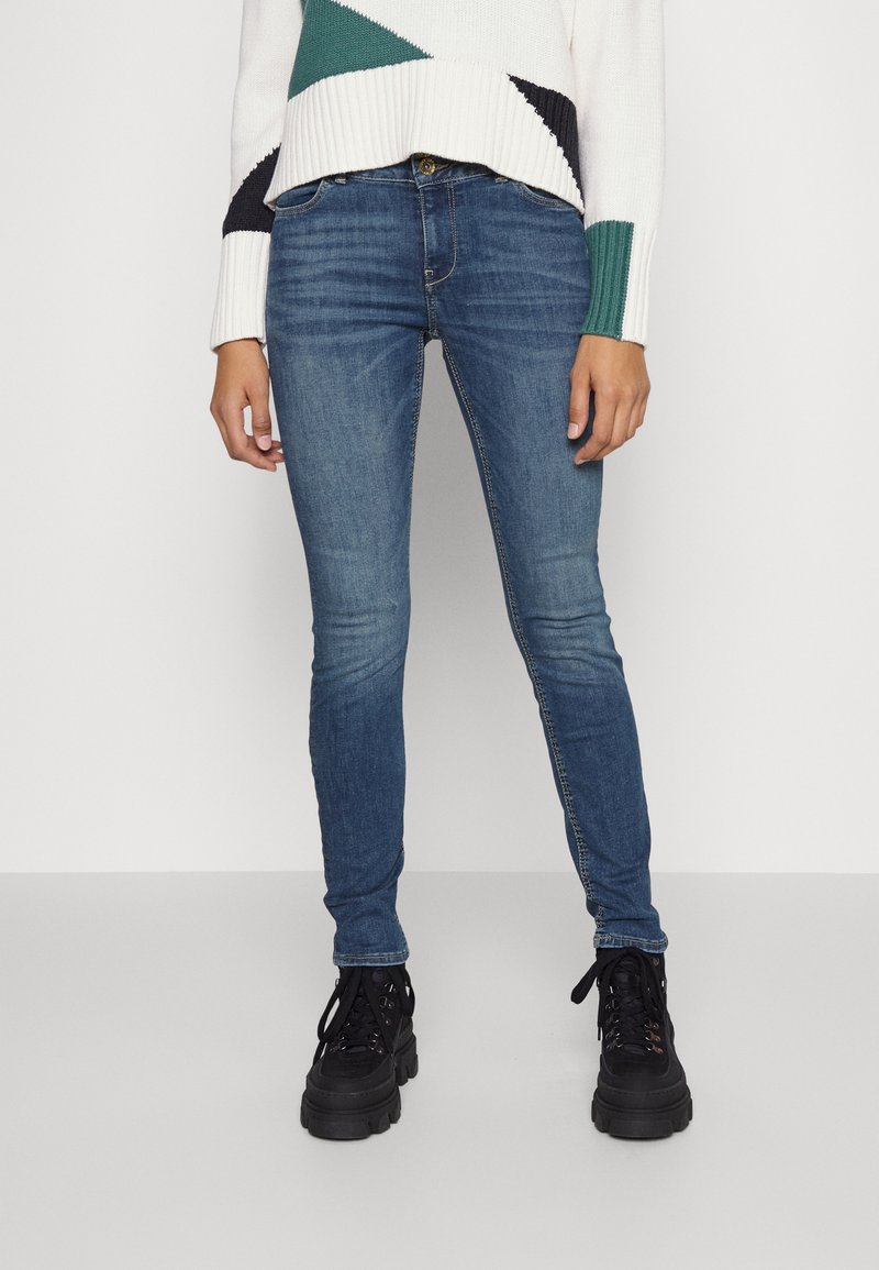 Femme portant un jean skinny bleu, des bottines noires épaisses à lacets, et un pull blanc avec des motifs géométriques verts et noirs.