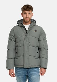 Homme portant une veste d'hiver grise rembourrée avec une capuche, une chemise blanche en dessous, et un jean bleu clair, se tenant contre un fond blanc.