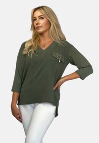 Oliwkowa bluzka z dekoltem w serek i rękawami 3/4, z kieszonką na piersiach i łańcuszkowym detalem. Miękka tekstura; luźny krój, zestawiona z białymi spodniami.