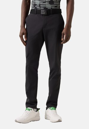 Mand iført sorte slim-fit bukser, hvide sneakers med grønne snørebånd og et sort bælte med en firkantet spænde.