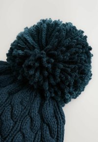 Bonnet tricoté en teal foncé avec un grand pompon, présentant un motif de câble texturé sur l'ensemble du corps. Matériau en mélange de laine douce.