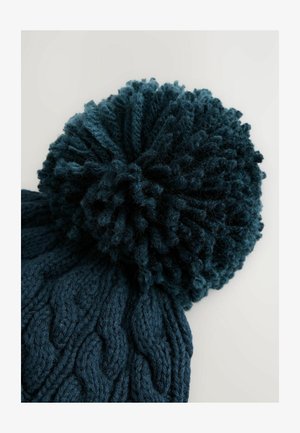 Pletená čepice v tmavě modré barvě s texturovaným vzorem kabelů a velkým, načechraným pom-pomem na vrchu. Materiál vypadá měkce a bujně.