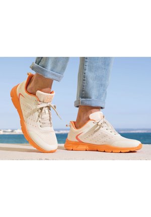 Persoon die lichtoranje en witte sneakers met veters draagt en opgerolde lichtblauwe jeans, staand bij een zee onder een heldere lucht.