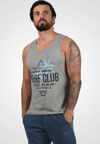 Sivi pamučni majica bez rukava s plavim i crnim grafičkim natpisom "Surf Club" i dizajnom palme. Uparen s tamnoplave trenirke.