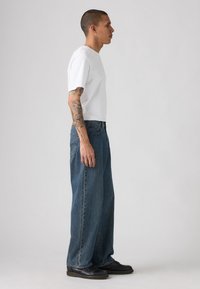 Hombre con camisa de manga corta blanca y jeans azules holgados, mostrando tatuajes detallados en los brazos, de pie en perfil contra un fondo claro y liso.