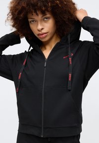 Svart zip-up hoodie med en figurnära design, med röd "HUGO" märkning på huvan och framsidan. Tillverkad av mjukt och slätt tyg med en känguruficka.