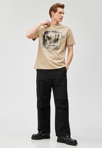 Camiseta gráfica beige con un diseño de calavera negra, combinada con pantalones cargo negros holgados y zapatos negros con suelas gruesas.