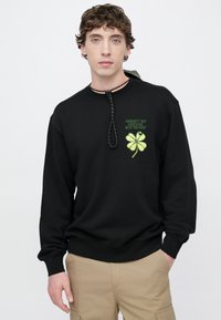 Felpa nera in pile con maniche lunghe, ricamo verde a trifoglio e testo sul davanti, coulisse nera regolabile al collo.