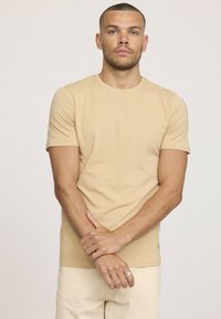Homme aux cheveux courts portant une chemise beige à manches courtes et un pantalon clair, se tenant les bras croisés devant un fond uni clair.