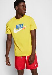Rumena bombažna majica s kratkimi rokavi ima velik modro-rdeč Nike znak, v kombinaciji z rdečimi kratkimi hlačami z belimi vrvicami in belim Nike znakom.