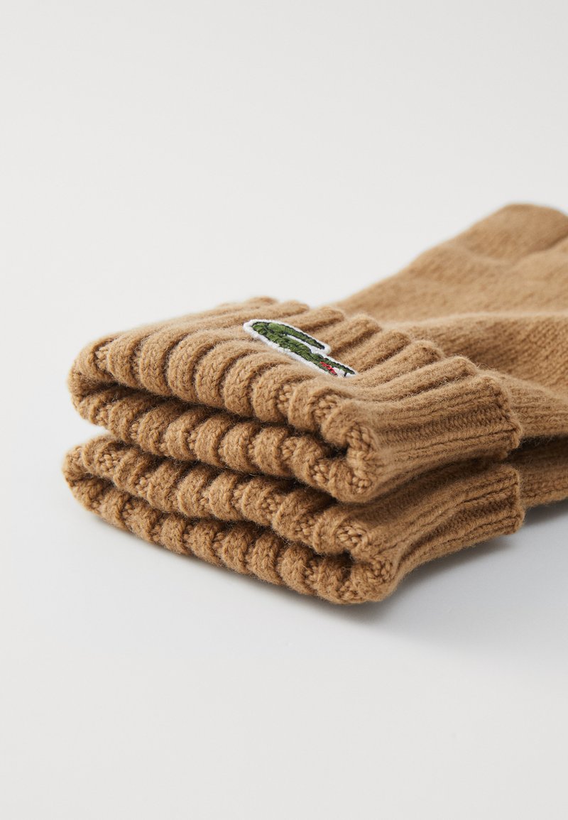 Brązowe, dziergane skarpetki ułożone starannie, z teksturą prążkowaną i małym, haftowanym zielonym logo aligatora na mankiecie.