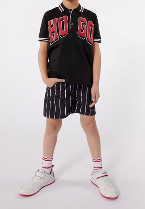 Black polo shirt with red "HUGO" lettering, striped black shorts, and white sneakers with red accents. White socks with red stripes visible.