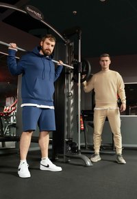 Due uomini in una palestra, uno con una felpa blu e pantaloni corti, l'altro con una felpa beige e pantaloni della tuta, entrambi con scarpe da ginnastica.