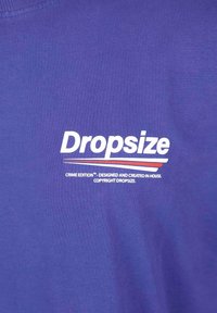 Lila T-Shirt mit dem Text "Dropsize" in Weiß, sowie "CRIME EDITION™" und zusätzlichen Branding-Details in weißen und roten Streifen.