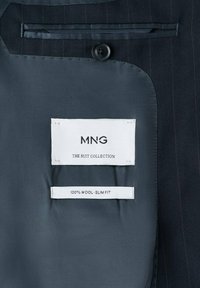 Indersiden af en mørk uldjakkesæt med en glat, grå foring. Mærkerne viser "MNG", "THE SUIT COLLECTION" og "100% ULD - SLIM FIT." Knappedetalje er synlig.