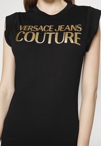 Svart t-shirt med kort ärm som har fet guldfolie-texten "VERSACE JEANS COUTURE" över framsidan. Mjuk tygkvalitet, figursydd design, med axelaccenter.
