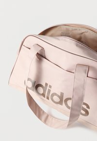 Ροζ υφασμάτινη τσάντα duffel με σατέν φινίρισμα, που διαθέτει ευδιάκριτο μεταλλικό χρυσό λογότυπο «adidas», διπλές λαβές και φερμουάρ στην κορυφή.