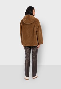 Braune Fleecejacke mit Kapuze, übergroßer Schnitt und strukturiertem Stoff, kombiniert mit braunen Lederhosen und hellen Schuhen.