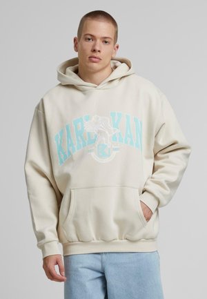 Sudadera beige con un bolsillo frontal. "KARL KAN" impreso en azul claro en el pecho, con un logo gráfico y puños acanalados.