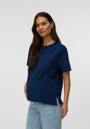 Zwangere vrouw met lang donker haar, gekleed in een marineblauw shirt met korte mouwen en lichtblauwe spijkerbroek tegen een effen lichte achtergrond.