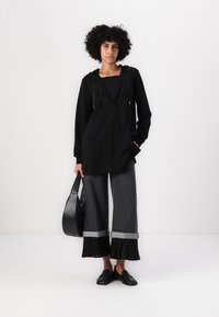 Felpa nera con cappuccio e dettagli in strass, abbinata a pantaloni grigi scuri a gamba larga con orlo plissettato. Modella che tiene una borsa nera.