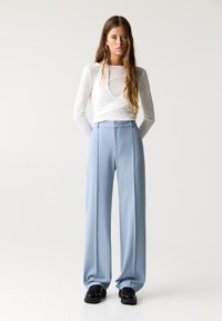 Weißes Langarmoberteil mit Wickeldesign, kombiniert mit hellblauen, weit geschnittenen Hosen mit glatter Textur und vorderen Falten. Schwarze Schuhe runden den Look ab.