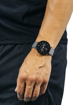 Orologio nero con quadrante rotondo, indicatori argentati e cinturino in tessuto grigio, indossato su una mano appoggiata su abbigliamento nero.