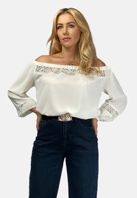 Biała bluzka bez ramiączek z koronkowym wykończeniem przy dekolcie i rękawach. Miękka tkanina i luźny krój, zestawiona z ciemnymi dżinsami z wysokim stanem.