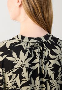 Bluse med blomsterdesign i svart stoff med lyse olivengrønne blomster, med rynket bakhals og myk tekstur.