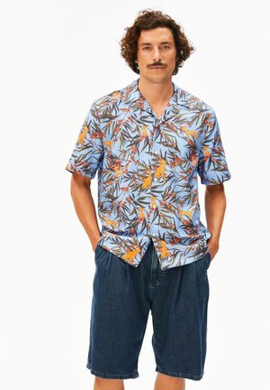 Mann mit lockigem Haar und Schnurrbart trägt ein blaues kurzärmeliges Hemd mit tropischem Muster und dunkle, lockere Jeansshorts, steht mit den Händen in den Taschen.