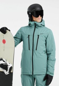 Mintgroene sneeuwjack met zwarte ritsen en accenten, voorzien van twee ritszakken en een capuchon, gecombineerd met een zwarte helm en snowboard.