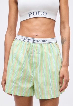 Femme portant un short boxer rayé vert de Polo Ralph Lauren et un crop top blanc avec le texte "POLO", montrant le ventre et un tatouage sur l'avant-bras.