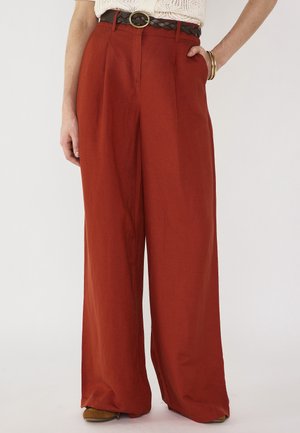 Pantaloni a gamba larga a vita alta di colore ruggine con tasche laterali, abbinati a una cintura intrecciata marrone scuro e braccialetti d'oro al polso.