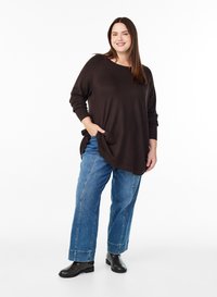 Ženska stoji z roko v žepu, oblečena v ohlapen temno rjav pulover, modre široke kavbojke in črne gležnjarje, na belem ozadju.