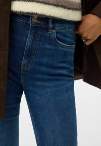 Sinised teksad, kitsas lõikega, kuldse õmblusega ja nööbiga kinnitatavad. Tekstuur on sile ja reitel on kerge pleekimine.