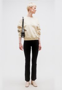 Άτομο με κοντά ξανθά μαλλιά, που φοράει μπεζ φούτερ, μαύρο παντελόνι, μπεζ μποτάκια μέχρι τον αστράγαλο και μαύρη τσάντα ώμου, κοιτάζοντας προς το πλάι.