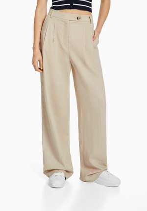 Pantalons beiges à taille haute et jambes larges avec boutons et plis, associés à des baskets blanches et un haut court à rayures noires et blanches.
