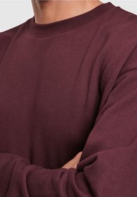 Primo piano di una persona che indossa un maglione girocollo bordeaux a maniche lunghe con le braccia incrociate in vita.