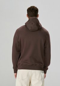 Brauner Kapuzenpullover mit weicher Textur, ausgestattet mit Kordelzug und gerippten Bündchen. Konzipiert für eine lockere Passform, getragen mit hellen Hosen.