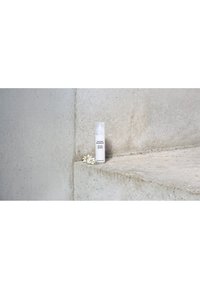 White Susanne Kaufmann skincare bottle on concrete step next to small white flowers, minimal background.
