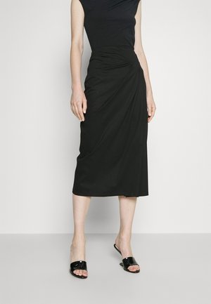 Kobieta ubrana w czarną sukienkę midi bez rękawów z detalem z marszczeniem po bokach oraz czarne sandały na obcasie z odkrytymi palcami, stojąca na jasnoszarej podłodze.