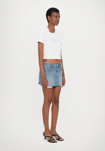 Tricou alb scurt cu logo-ul "KSUBI", fustă mini din denim cu tiv destrămat și sandale negre cu toc subțire.
