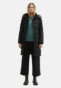 Schwarze Daunenjacke mit einer fütterten Kapuze, vorderem Reißverschluss und großen gesteppten Abschnitten, getragen über einem türkisfarbenen Pullover und schwarzen weit geschnittenen Hosen.