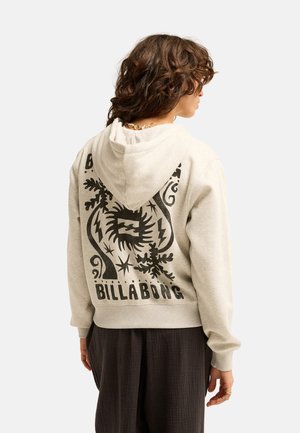 Osoba z kręconymi włosami, ubrana w beżową bluzę z kapturem z dużą czarną grafiką i tekstem "BILLABONG" na plecach, w połączeniu z ciemnymi, luźnymi spodniami.
