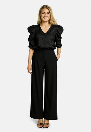 Uśmiechnięta kobieta o blond włosach, ubrana w czarną bluzkę z bufkami, szerokie czarne spodnie oraz czarne sandały na paski, stoi na tle białego tła.