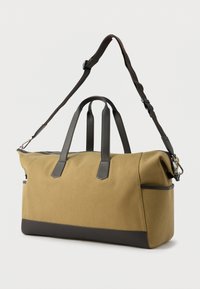 Μεγάλη ορθογώνια τσάντα duffel από καμβά σε ανοιχτό καφέ χρώμα με μαύρες δερμάτινες λαβές, μαύρο περίγραμμα στην κάτω πλευρά και αποσπώμενο ρυθμιζόμενο λουρί ώμου, σε λευκό φόντο.